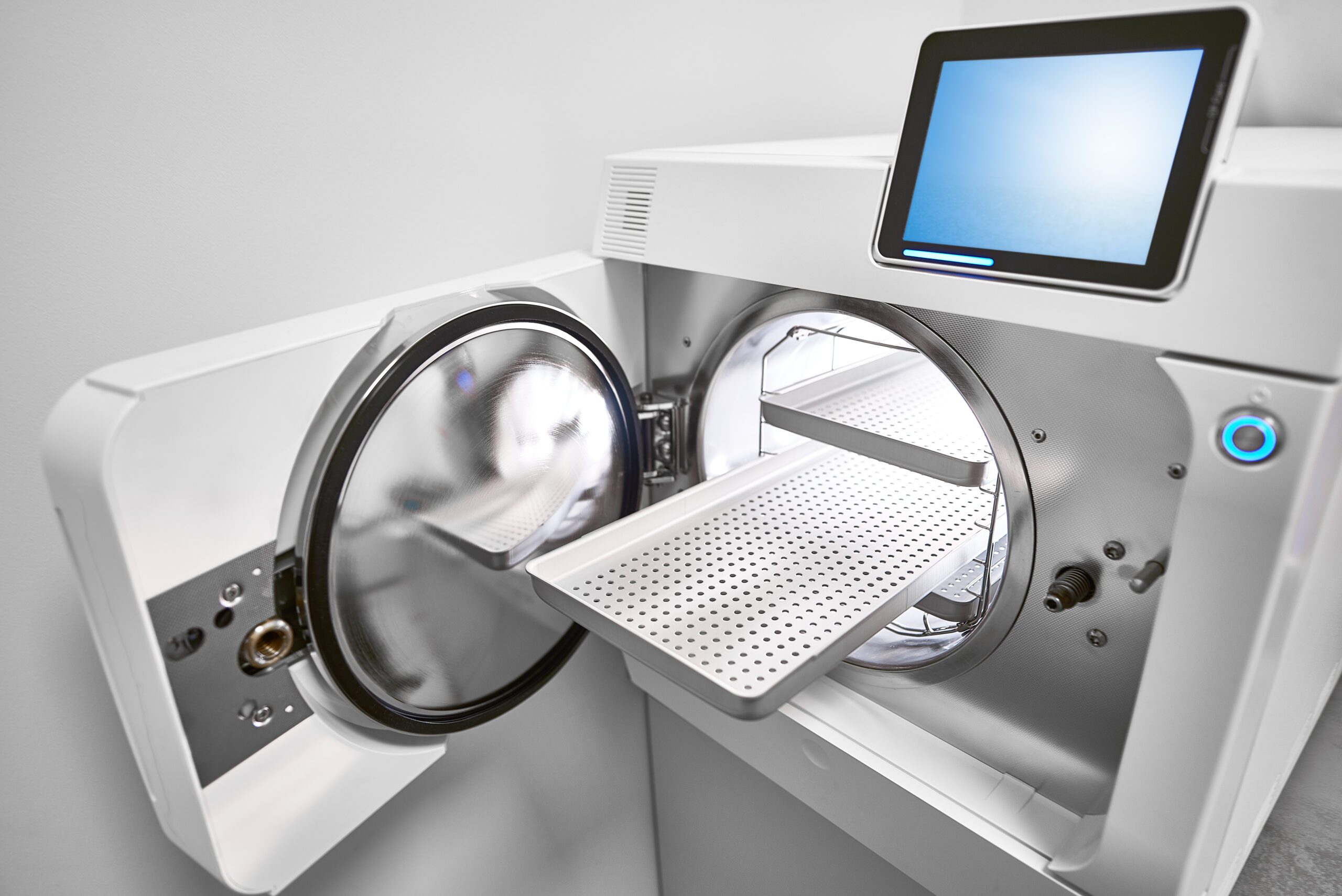 An autoclave machine has an open door with one of the three trays pulled out and empty. The autoclave has a digital screen.