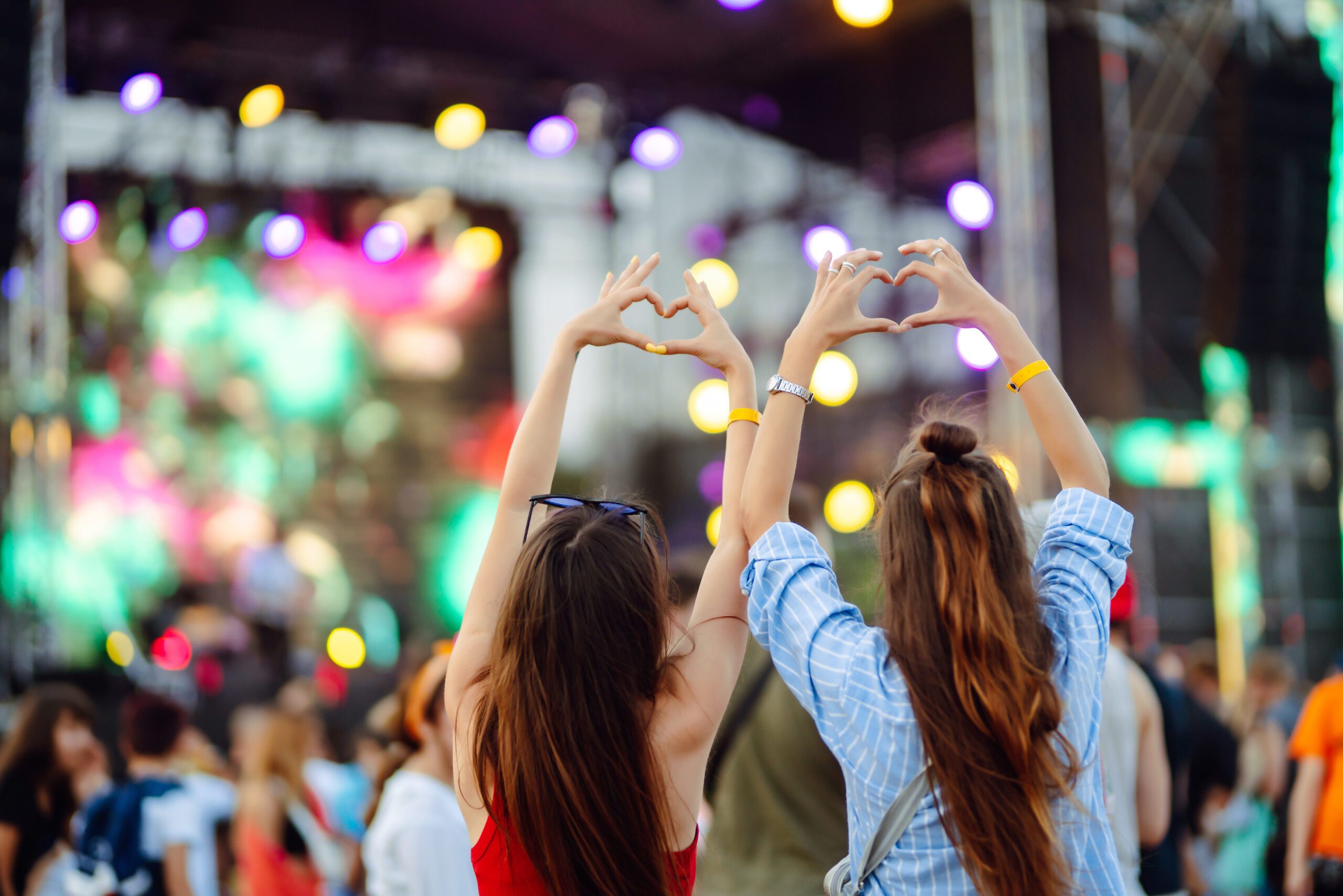 Two girls with their hands above their heads, making hearts with their fingers. they're watching a brightly colored stage