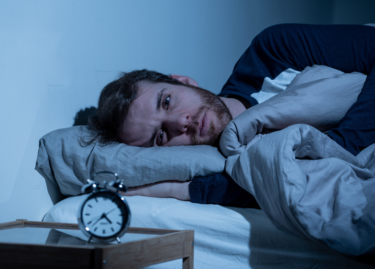 A distressed man laying on his side in bed. The lights in the room are off and there's a small alarm clock on his bedside table.