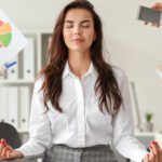 Home 10 A young businesswoman sits on a desk with her legs crossed and eyes closed. Arms are holding out paperwork, pens, and a phone.