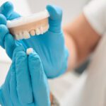 Home 9 A dental professional wearing blue latex gloves holds a fake crown in one hand and an impression of a top jaw in the other.