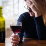 A woman rests one hand on her forehead while grasping a wine glass full of red liquid with the other.