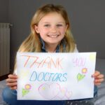 A young girl sits on a bed with a "Thank You, Doctors!" drawing. A dresser and a flower vase are in the background.