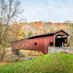 A red, covered bridge goes over a small creek. Mature trees with red, orange, and yellow leaves are in the background.