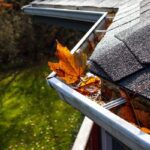 A close-up view shows several orange leaves sitting in a home’s silver gutters next to black shingles.