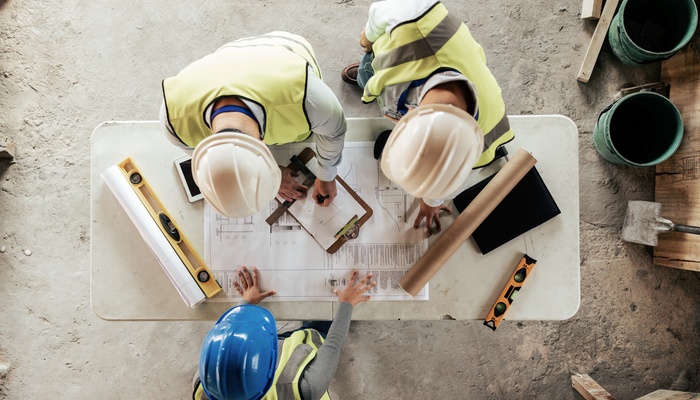 A construction team, wearing reflective gear and hard hats, is reviewing architecture sketches for a renovation project.
