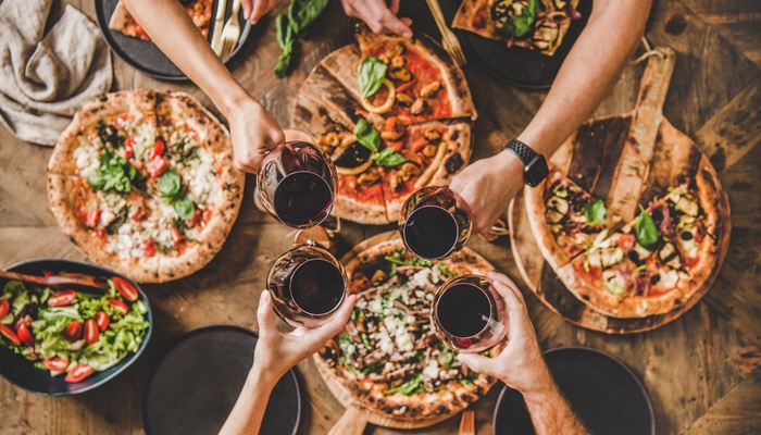 Friends reaching across a table loaded with individual pizzas, with each person holding a glass of red wine to toast.