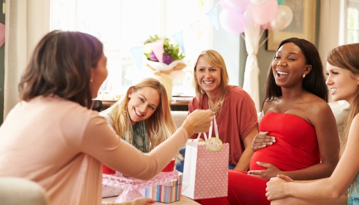 Five women sit together at a baby shower. One woman is giving a gift to the pregnant woman at the party.