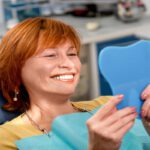 Home 7 A woman in a yellow shirt lies in a blue dental chair, smiling and holding a blue tooth-shaped mirror.