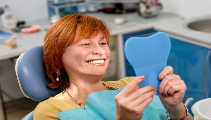 A woman in a yellow shirt lies in a blue dental chair, smiling and holding a blue tooth-shaped mirror.