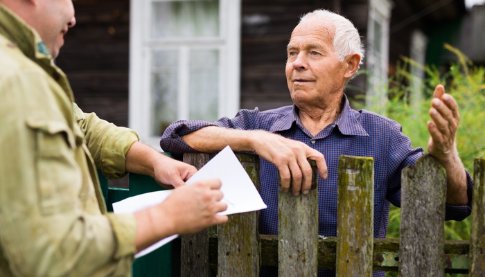 An older man is communicating with an insurance company rep while standing at the fence of his house.