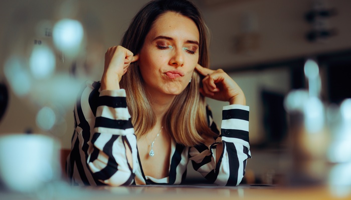 A woman in a striped shirt sits at a restaurant table with her eyes closed and plugs her ears with her fingers.