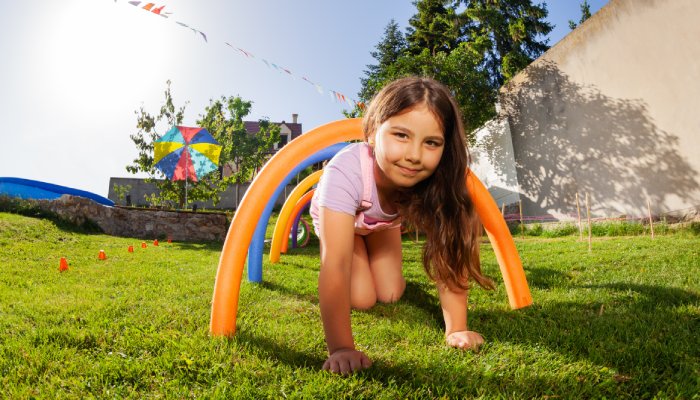 Home 17 A brown-haired girl crawls on grass under an orange pool noodle arch, with four colorful arches lined up behind her.