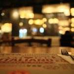 A fork and knife rest on a napkin placed over a menu on a wooden table with blurred lights and diners in the background.