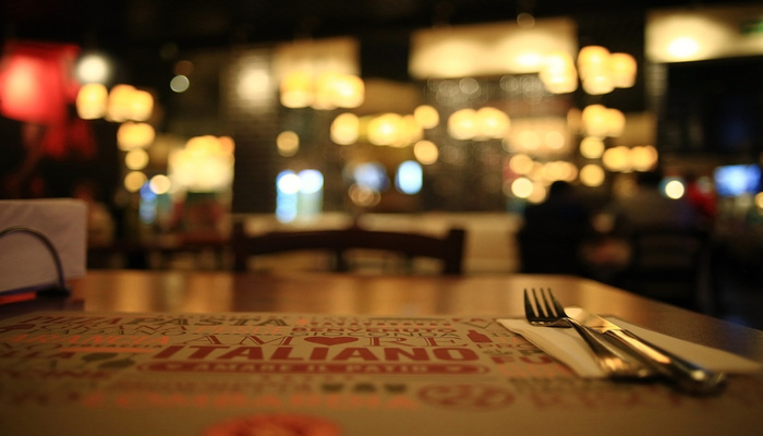 A fork and knife rest on a napkin placed over a menu on a wooden table with blurred lights and diners in the background.