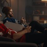 Two friends, wearing blue and red football jerseys, sit on a couch in a garage, playing video games.