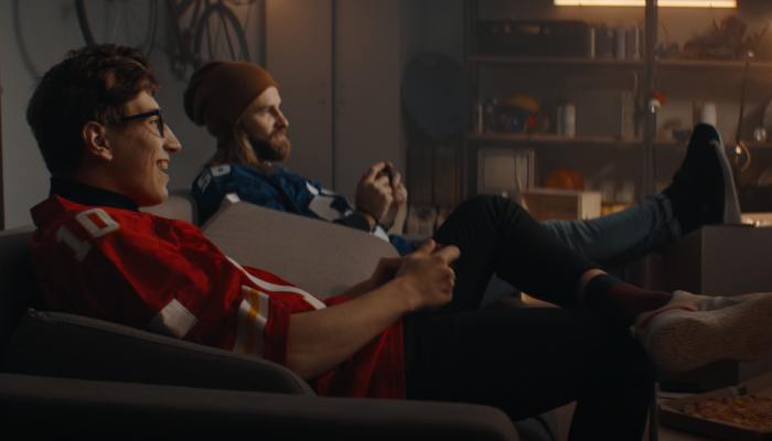 Two friends, wearing blue and red football jerseys, sit on a couch in a garage, playing video games.