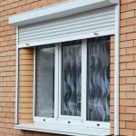 A white rolling shutter is over a white window on a red-colored brick home. Blue and gray curtains hang behind the window.
