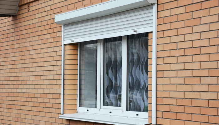A white rolling shutter is over a white window on a red-colored brick home. Blue and gray curtains hang behind the window.