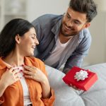 A man surprises a woman sitting on a gray couch with a gift wrapped in red wrapping paper and a white bow.