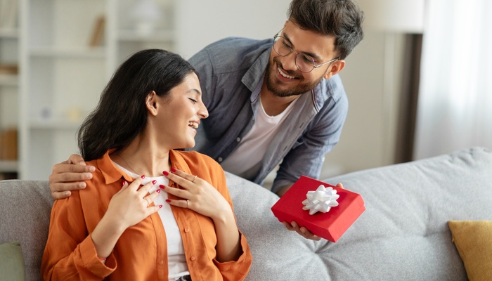 A man surprises a woman sitting on a gray couch with a gift wrapped in red wrapping paper and a white bow.