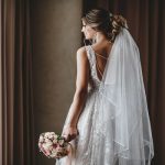 A bride in a white, sleeveless wedding gown with intricate back details and a long veil holds a bouquet.
