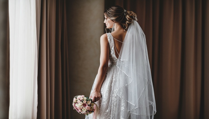 A bride in a white, sleeveless wedding gown with intricate back details and a long veil holds a bouquet.
