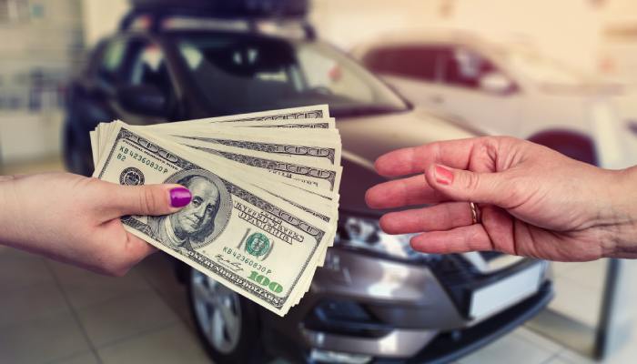 One hand is holding out a stack of bills, while another hand is reaching out to grab it. In the background there is a car.