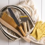 Various beekeeping tools like a beekeeping suit, a brush, and scrapers lay flat on a white wooden table.