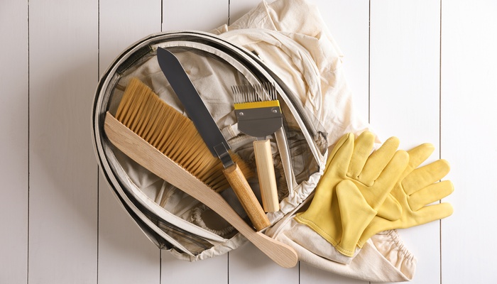Various beekeeping tools like a beekeeping suit, a brush, and scrapers lay flat on a white wooden table.