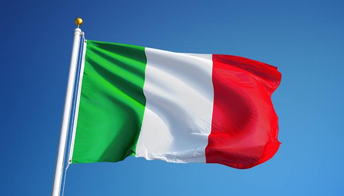 A green, white, and read Italian flag flies high in the sky on a white flagpole. There is a clear blue sky in the background.