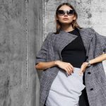 A young woman wearing sunglasses, a plaid trench coat, and a grey skirt poses outdoors by a concrete wall.