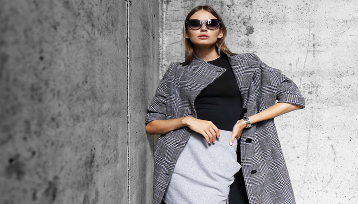 A young woman wearing sunglasses, a plaid trench coat, and a grey skirt poses outdoors by a concrete wall.