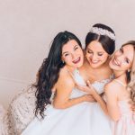 Three women, a bride and two bridesmaids, smile and hug in a light-colored room with a plain wall behind them.