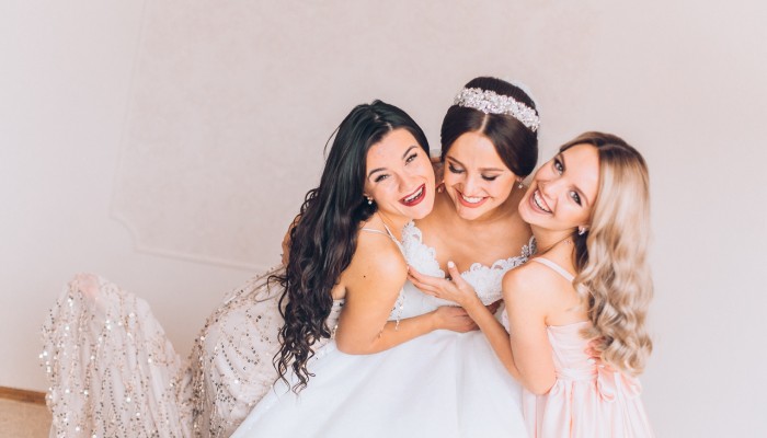 Three women, a bride and two bridesmaids, smile and hug in a light-colored room with a plain wall behind them.