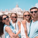 A family of four, wearing sunglasses, posing and smiling in front of St. Peter's Church in the Vatican, Italy.