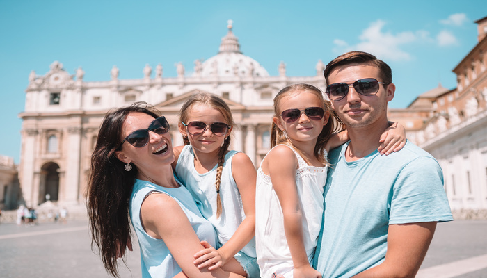 A family of four, wearing sunglasses, posing and smiling in front of St. Peter's Church in the Vatican, Italy.