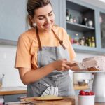 A young woman smiles as she puts whipped icing and red raspberries on a freshly baked cake in her kitchen.