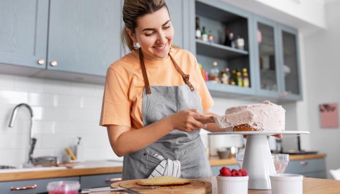A young woman smiles as she puts whipped icing and red raspberries on a freshly baked cake in her kitchen.