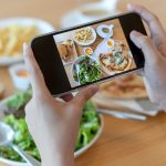 A table has a salad, a pizza, pasta, and a cup of coffee. A person holds a phone horizontally and takes a photo.