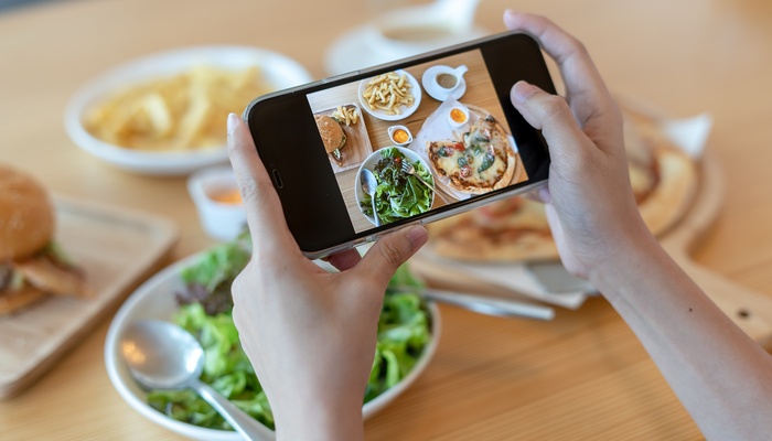 A table has a salad, a pizza, pasta, and a cup of coffee. A person holds a phone horizontally and takes a photo.