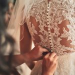Close-up of a bridesmaid fastening the back of a bride's intricate white wedding dress, with a sheer veil visible.