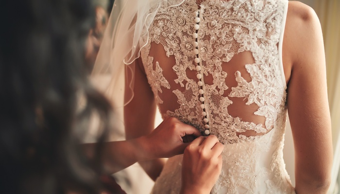 Close-up of a bridesmaid fastening the back of a bride's intricate white wedding dress, with a sheer veil visible.