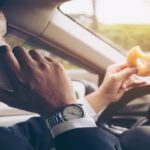 A man is driving a car while he is talking on the phone and holding a bagel in his left hand.