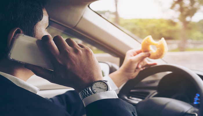 A man is driving a car while he is talking on the phone and holding a bagel in his left hand.