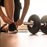 A person tying their workout shoes on at home. A water bottle and dumbbell are sitting on the floor.