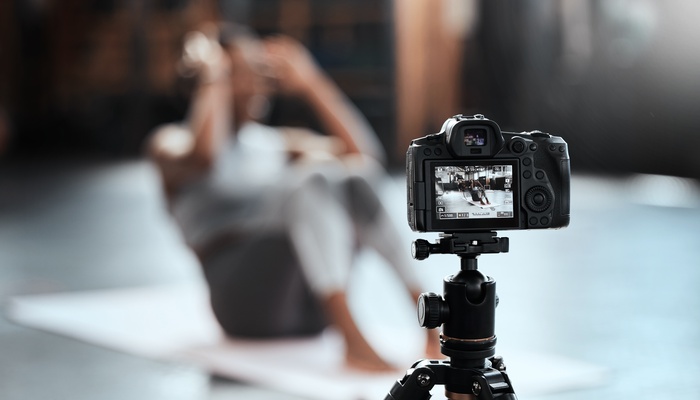 A close-up view shows a black, high-end digital camera sitting on a black tripod, taking a photo of a person doing a sit-up.